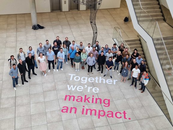 Eine Gruppe Menschen steht zusammen und schaut nach oben in die Kamera.Daneben der Schriftzug: Together we're making an impact.