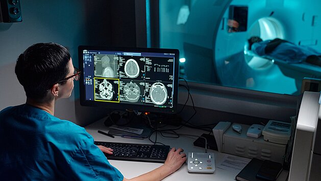 Medical professional reviewing patient's MRI images on computer monitor with patient in MRI machine in background.